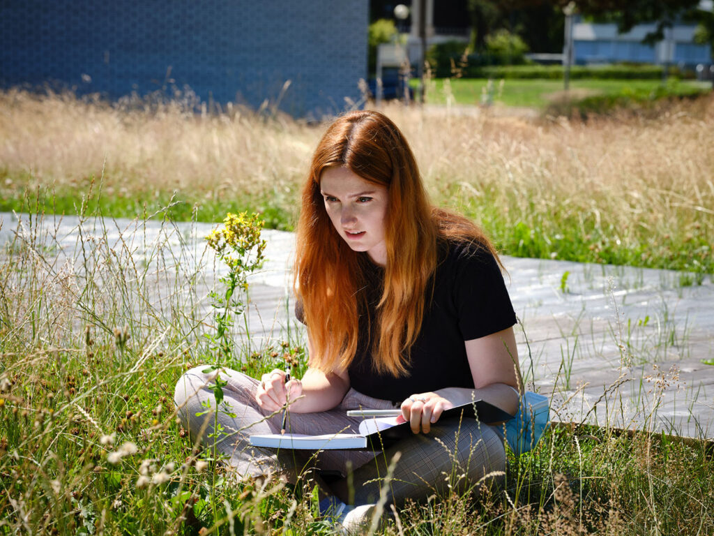 Woman sitting cross legged outside, sketichng a flower in a notebook.