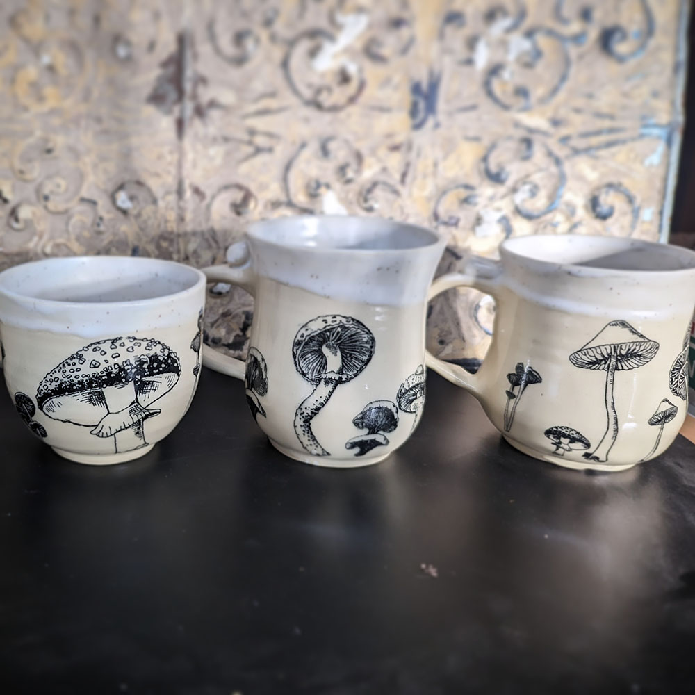 Three beige cereamic mugs with black mushrooms painted on them.