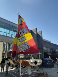 A robotic sailing vessel is set on land outside of a university building on planks