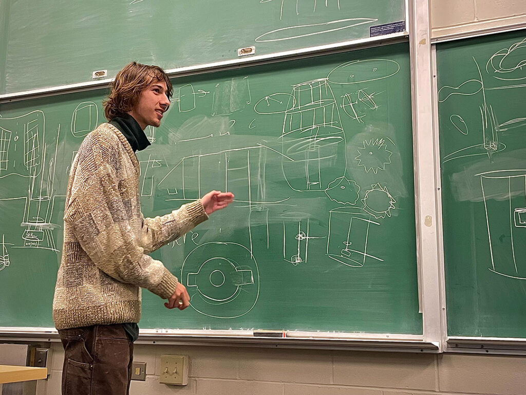 Charting New Waters_IMG_1866 A man stands at a chalkboard with shapes on it and looks off intently.