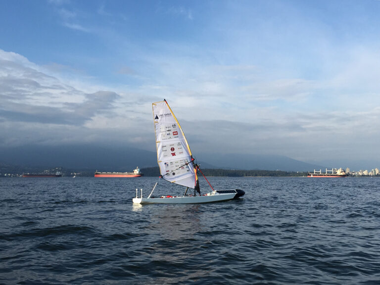 A robotic sailing vessel is out on water with the words "UBC Sailbot" on the sail
