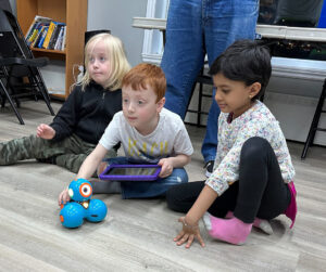 Three young children are seated on the floor, intently focused on a small blue and orange robot, known as Dash and Dots. One boy in a white t-shirt holds a tablet, controlling the robot, while a child in a black hoodie and a girl in a multicolored sweater watch closely.