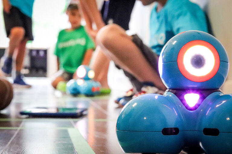 A close up shot of a Dash and Dots robot, comprising three blue spheres and wheels, with children playing the background