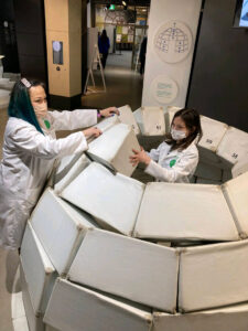 Two people work together to build a model igloo at a science fair, focusing on assembling white panels.