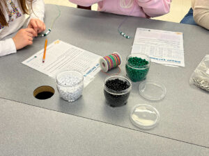 A close-up of a tabletop where two children are participating in a binary encoding activity. They are threading white, green, and black beads onto strings to represent binary code. Containers of beads, along with rolls of string, are placed on the table. A worksheet with binary encoding instructions and a pencil are also visible, guiding the activity. The children are focused on creating their bead sequences, engaging in the hands-on learning exercise.
