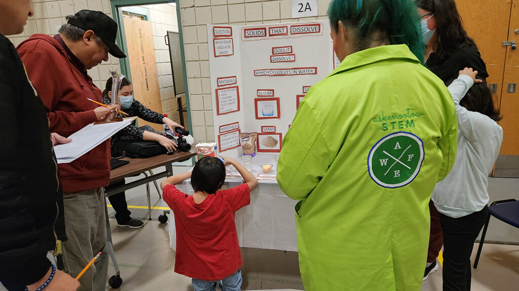 Science Fair_Impact A person attends a science fair, standing in front of a project display about Indigenous knowledge and STEM