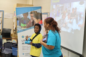 A woman and a young boy are giving a demonstration at a school event. The boy holds a blue robot, while the woman holds a tablet. Both are wearing bright yellow and blue shirts with the Pinnguaq logo. In the background, a projector displays an image of children working on laptops, and a banner for Lindsay Makerspace is visible.
