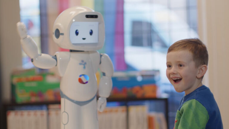A young boy with a joyful expression interacts with a small humanoid robot in a bright, colorful room. The robot, with a friendly face on its screen, waves its hand while standing next to the child. Shelves filled with books and educational materials are visible in the background, giving the scene a playful, learning environment feel.
