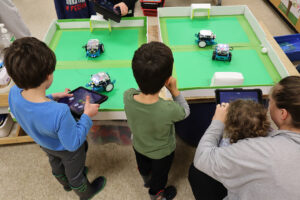 Several young children are gathered around a table where two small soccer fields are set up. Each field has miniature robots, controlled by tablets held by the children. The robots are positioned to play a soccer game, with small goals on either end of the fields. One child looks on while others interact with their tablets, programming or directing the robots. An adult helps a young child manage a tablet, engaging them in the activity.