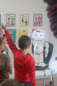 A young boy in a red hoodie raises his hand while facing a small, white humanoid robot in a classroom setting. The robot is raising one of its arms as well, engaging with the children. Behind the boy, other children are also watching and participating in the interaction.