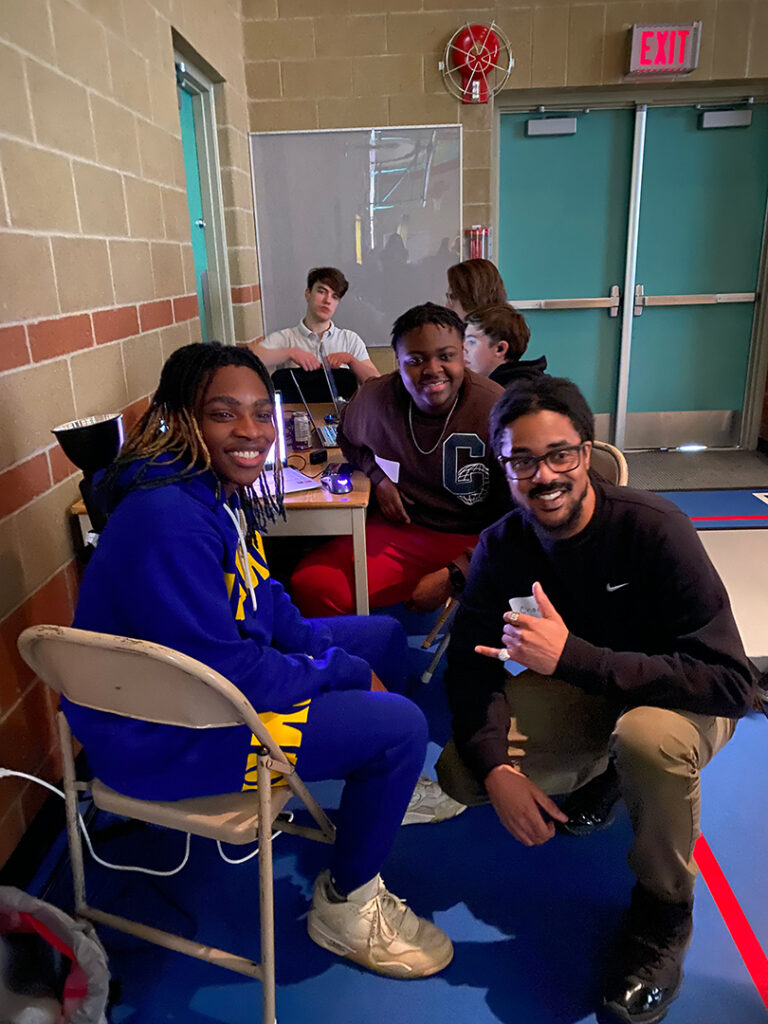 Students posing_Your Voice Is Power Three students are seated at a table in a room with brick walls and double doors. They are smiling and posing with Chad Allsopp, the Your Voice is Power hip-hop consultant, who is crouching in front of them. The setting appears to be a workshop or classroom, with laptops and other materials on the table behind them. The atmosphere is relaxed and collaborative.