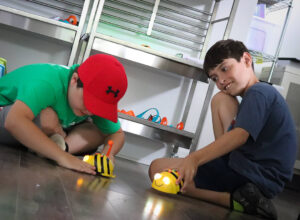 Two boys sit on the floor, playing with yellow and black striped robots designed to look like bees, known as Bee-Bots. One boy, wearing a green shirt and a red hat, is focused on interacting with his Bee-Bot, while the other, in a blue t-shirt, smiles as he operates his own. The scene takes place in a classroom.