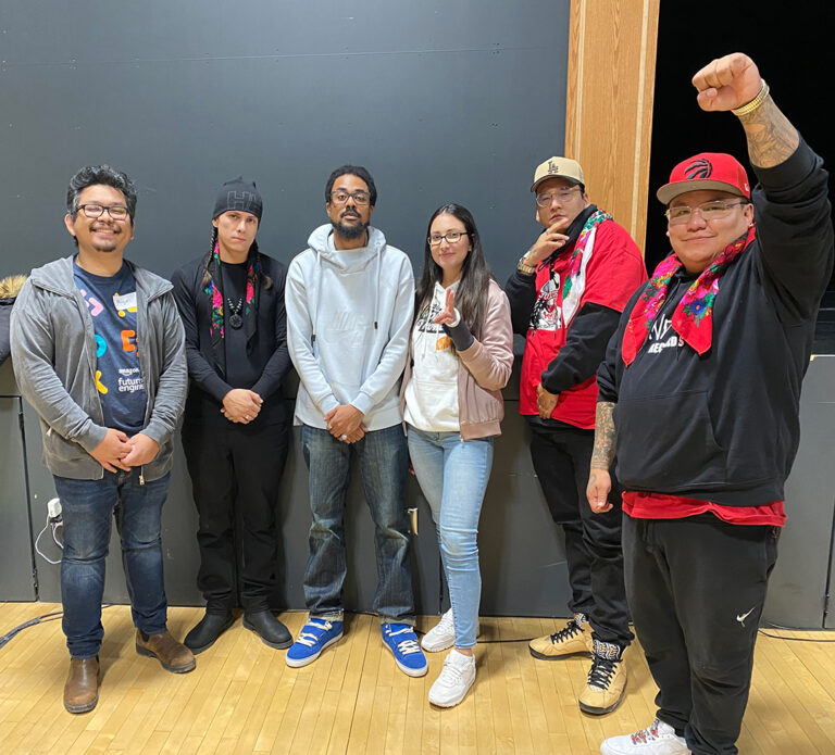 A group of six individuals standing side by side in a well-lit indoor space. They are casually dressed, smiling, and posing together. The person on the far right is raising a fist in the air. The group includes two women and four men. The background features a gray wall and a wooden frame on the right side.