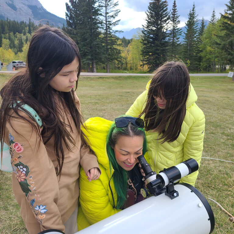 A woman and two children stand outdoors in a grassy area surrounded by trees and mountains. The woman looks through a telescope while the children watch. The scene is part of a STEM event in a natural setting, with the group engaged in astronomy activities.